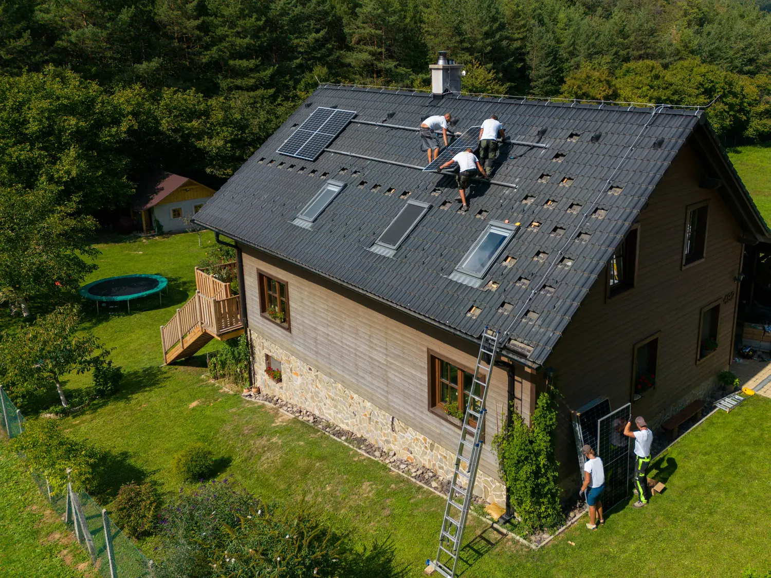 Blick von oben auf ein Wohnhaus, auf dessen Dach drei Monteure die Unterkonstruktion für die Solarmodule setzen