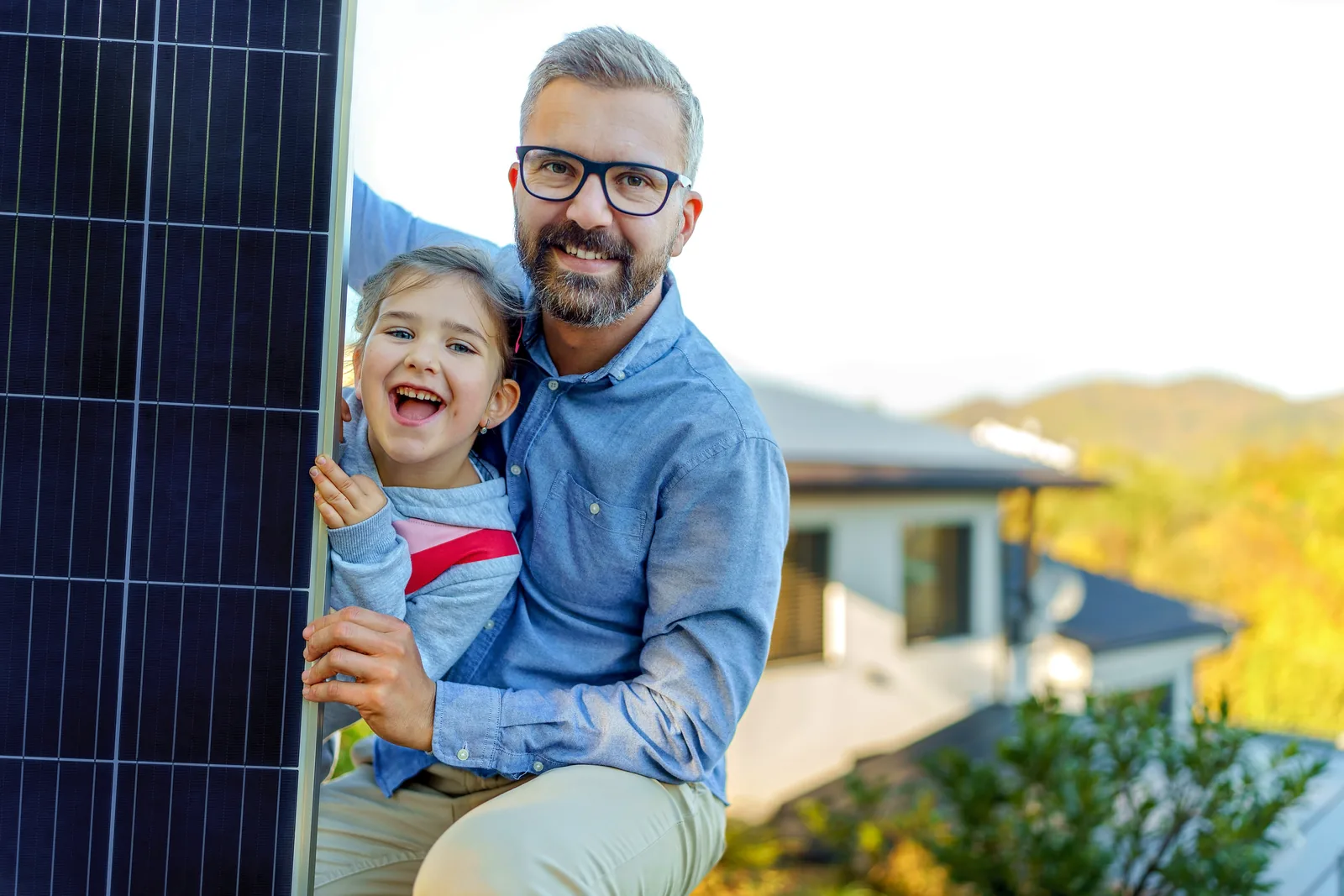 Vater und Tochter stehen lachend neben einem Solarmodul vor ihrem Haus